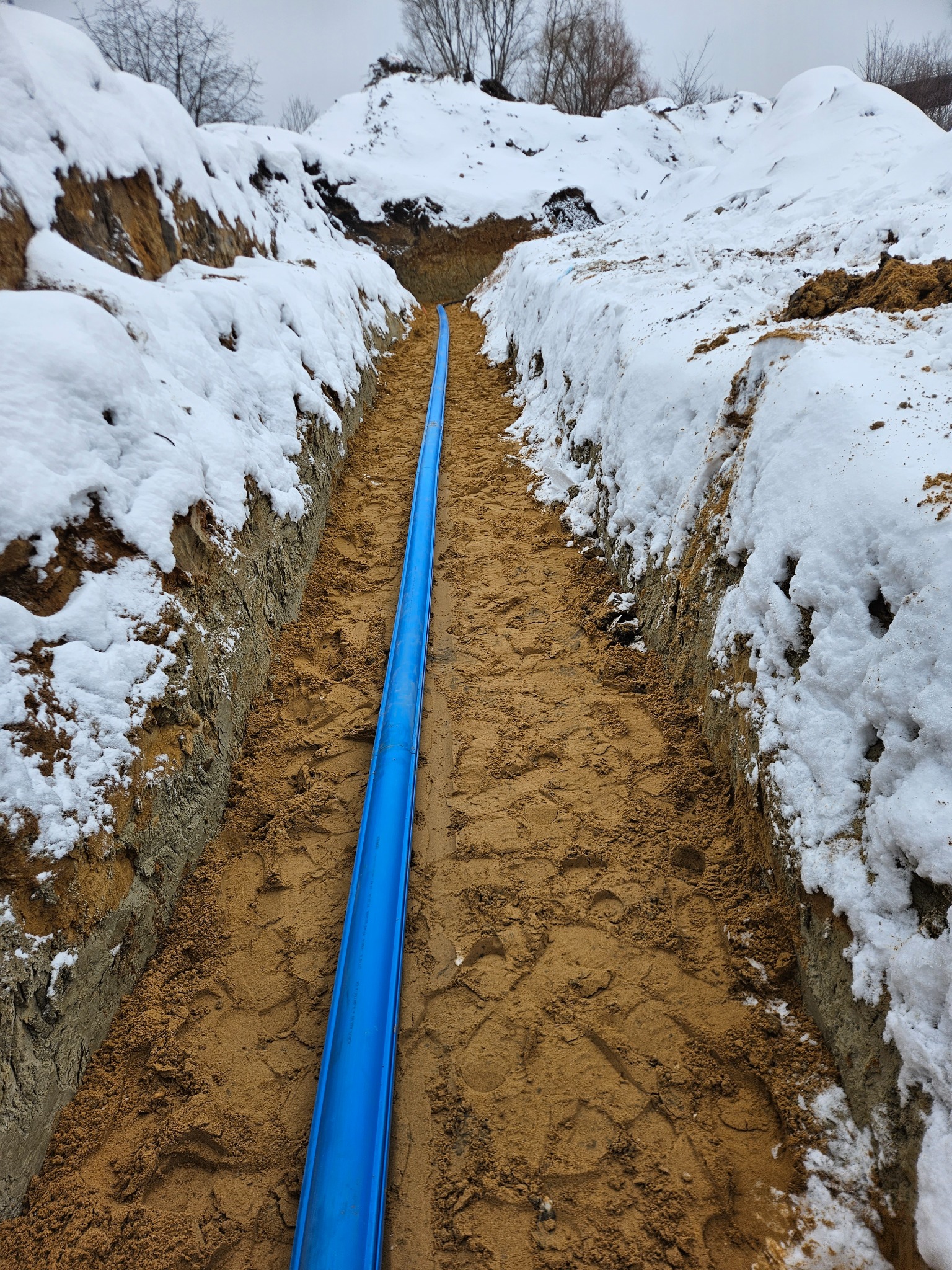 Niebieska rura w wykopie zasypanym piaskiem, obok ściany wykopu pokryte śniegiem, zimowy krajobraz.