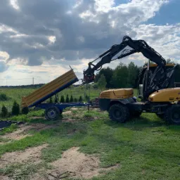Ż&oacute;łta koparka Mecalac z ramieniem podnosi przyczepę z ż&oacute;łtą skrzynią ładunkową na tle zielonego pola i pochmurnego nieba.