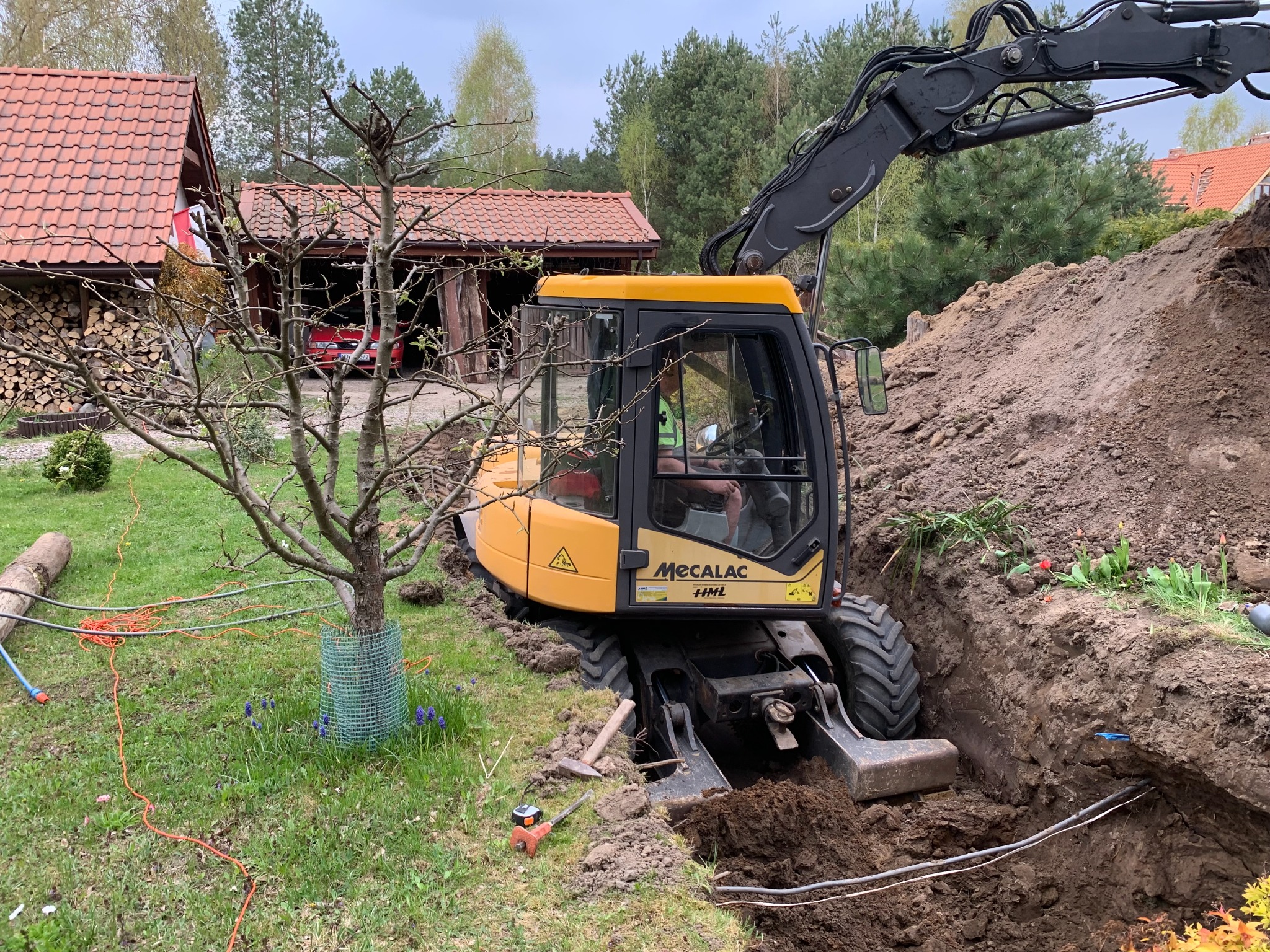 Żółta koparka Mecalac HML wykonuje wykop w ziemi, widoczne rury w wykopie, drzewo z zabezpieczeniem pnia, dom z czerwonym dachem w tle.