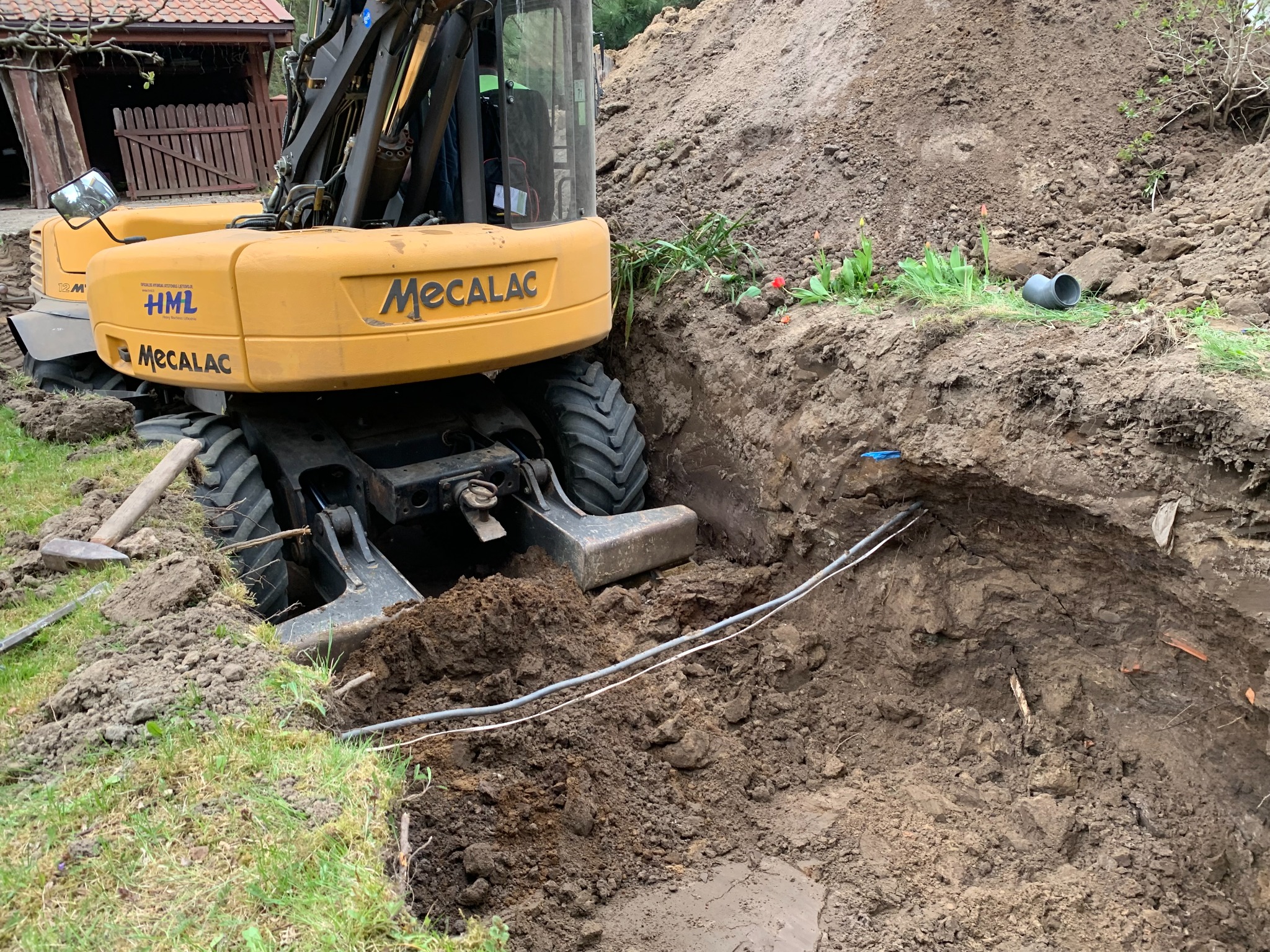 Żółta koparka Mecalac częściowo zagłębiona w wykopie pod instalacje, widoczne kable w ziemi, fragment rury PCV i pryzma ziemi.