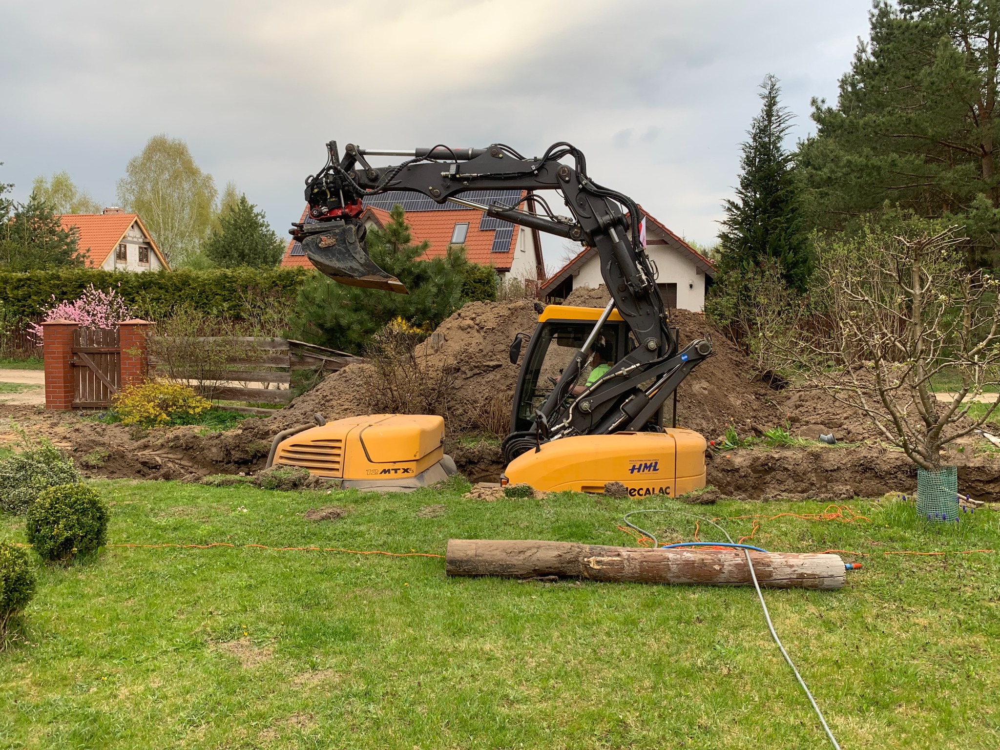 Żółta koparka HML Decalac w trakcie prac ziemnych na trawniku, widoczny wykop, fragment domu z panelami słonecznymi w tle.