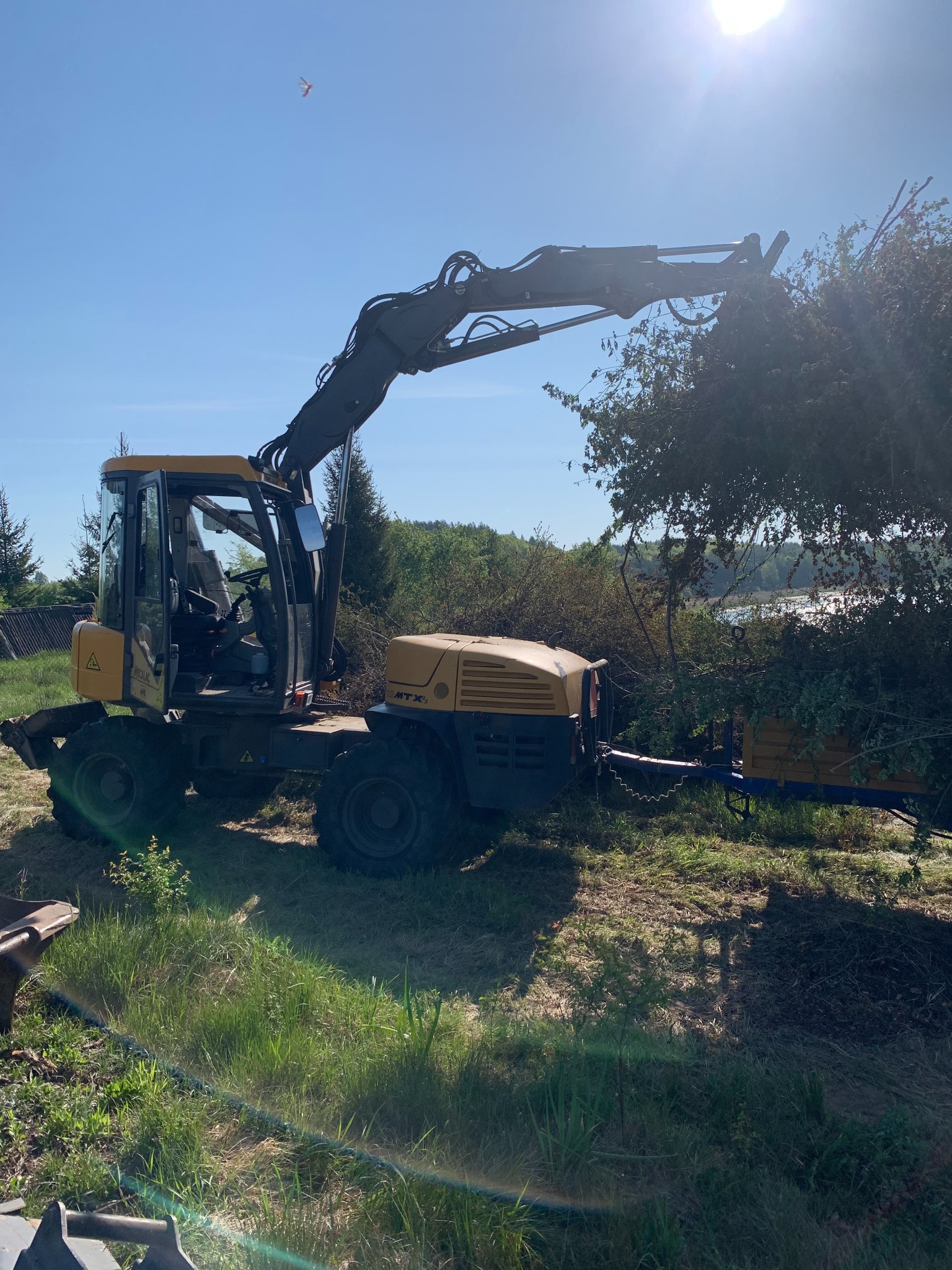 Żółta maszyna z ramieniem chwytającym gałęzie drzew, przyczepka z pojemnikiem na zrębki, w tle błękitne niebo i drzewa.