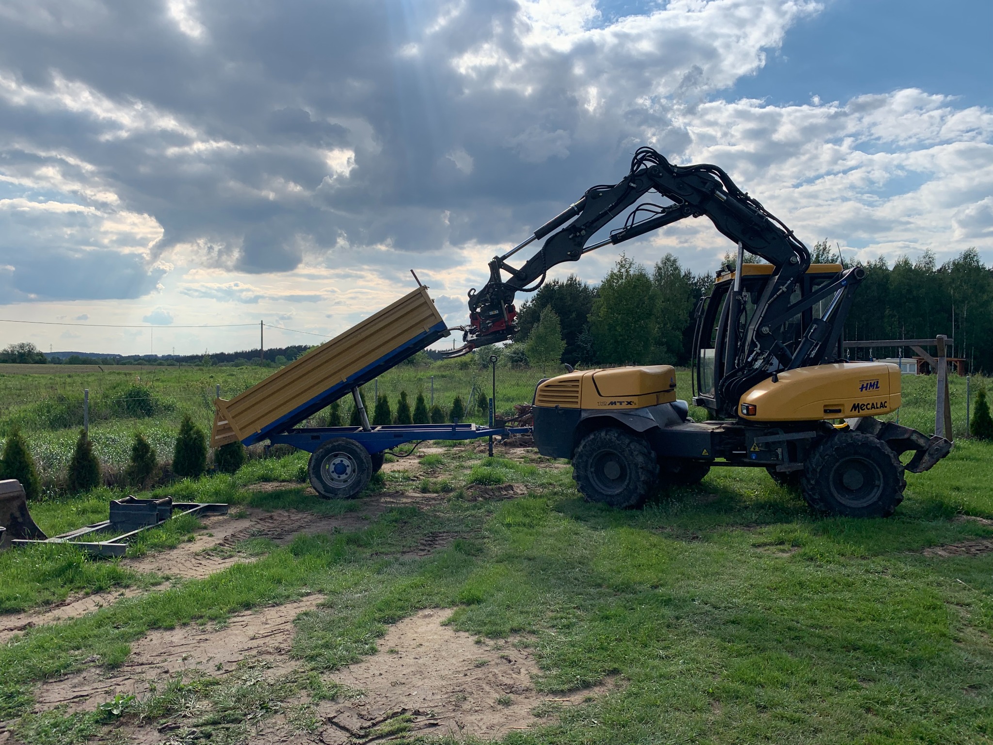 Żółta koparka Mecalac z ramieniem podnosi przyczepę z żółtą skrzynią ładunkową na tle zielonego pola i pochmurnego nieba.