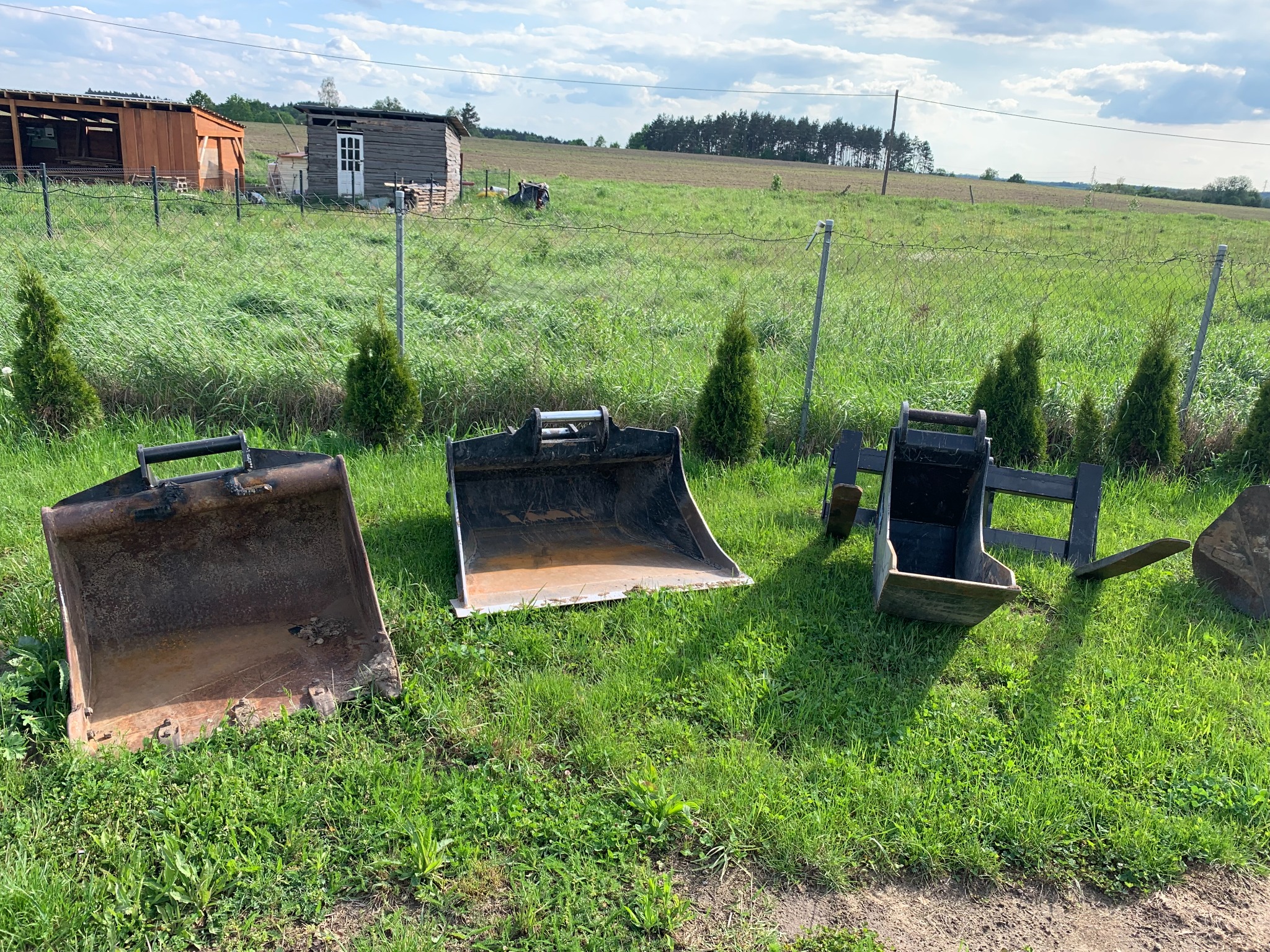 Trzy różne łyżki koparkowe ustawione na trawie przed polem i budynkami gospodarczymi, widoczne ślady użytkowania, zróżnicowane rozmiary i konstrukcje.