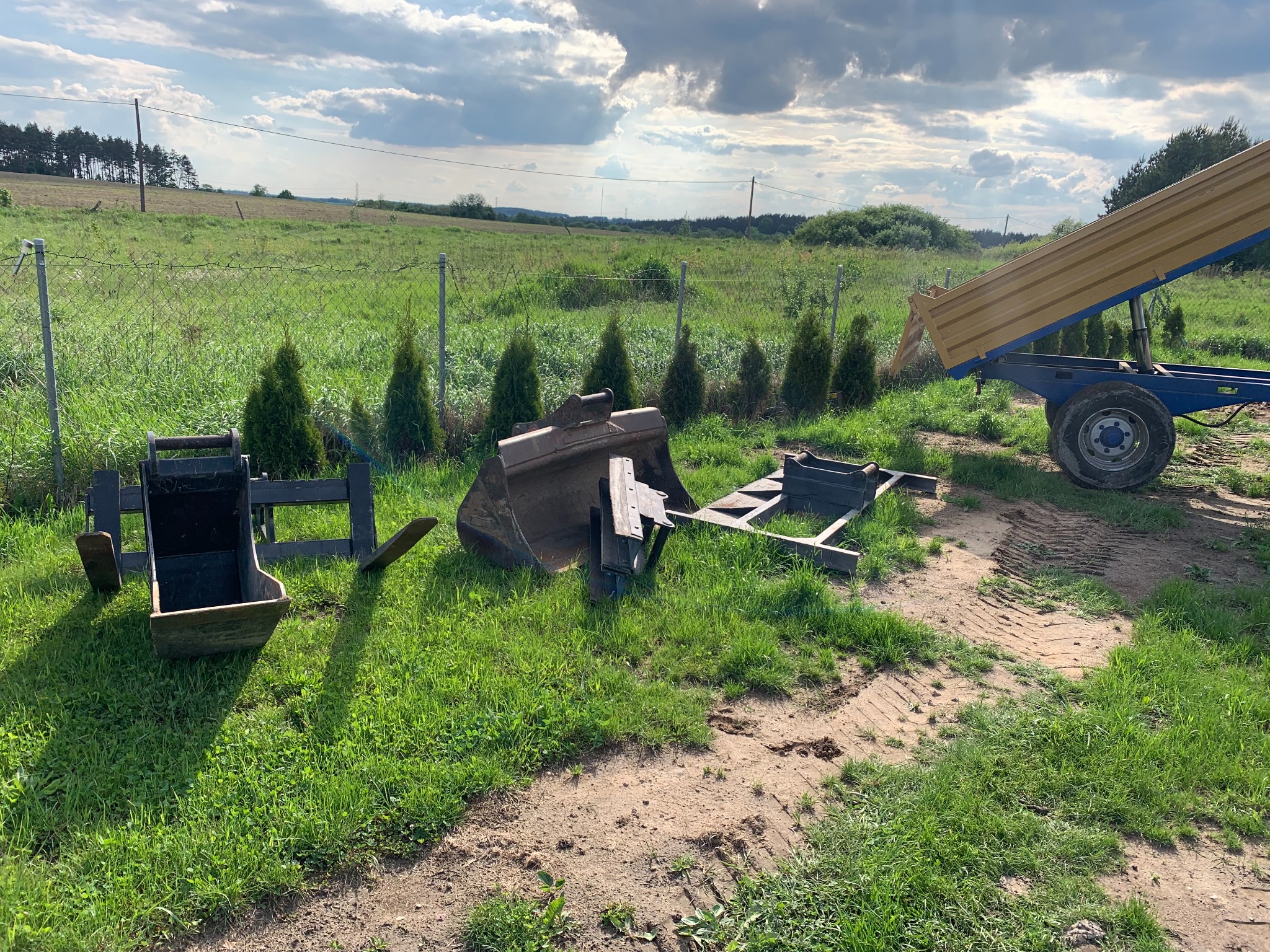 Na trawie leżą elementy koparki i przyczepa wywrotka z podniesioną skrzynią ładunkową, w tle ogrodzenie i pole.
