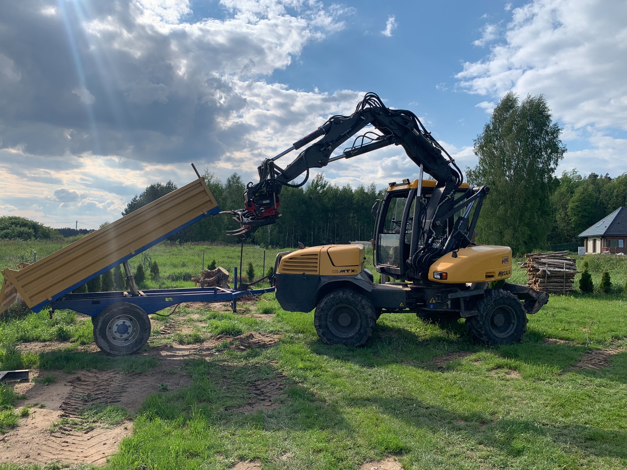 Żółta maszyna wielofunkcyjna z przyczepą na trawiastym terenie, chwytak na wysięgniku, w tle drzewa i dom.