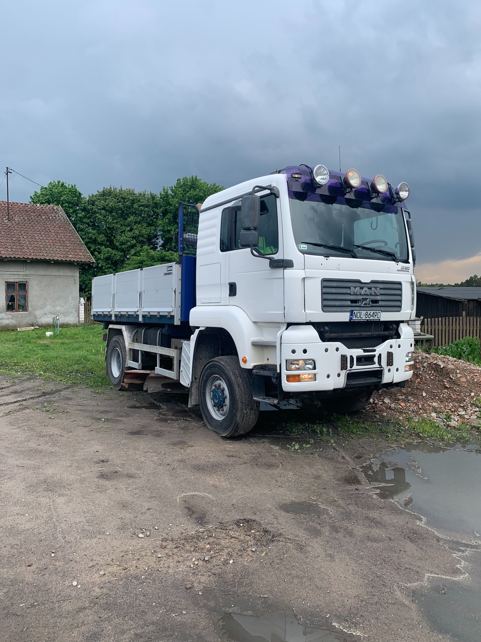 Biała ciężarówka MAN z platformą załadunkową stoi na błotnistym terenie w Barczewie, z widoczną stertą gruzu i kałużą; pochmurne niebo w tle.