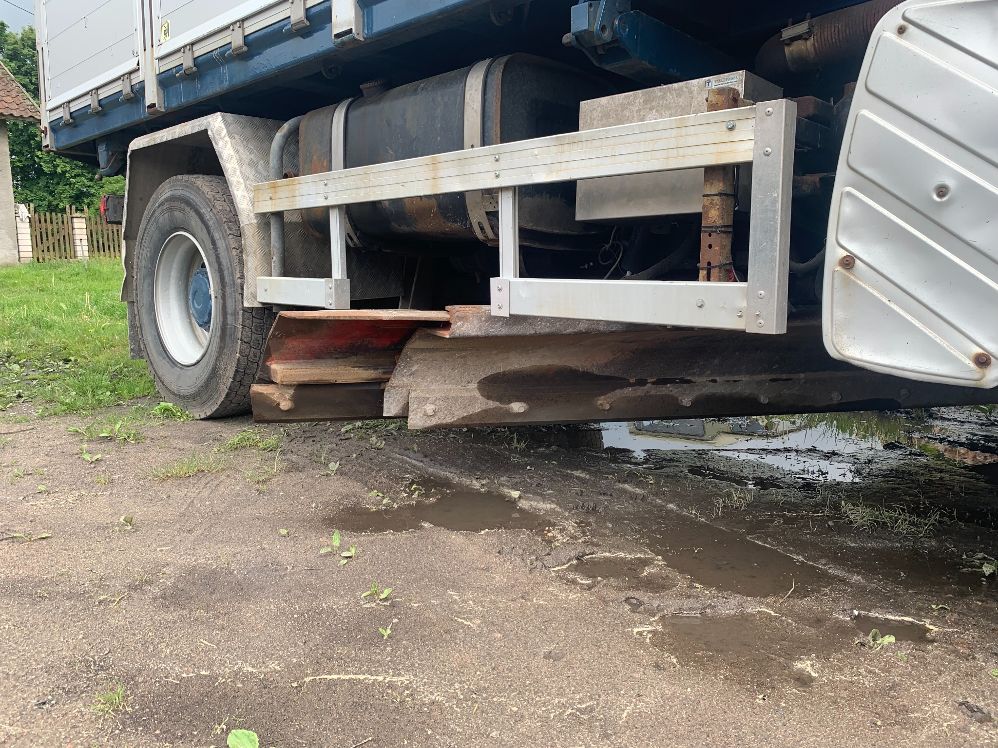 Fragment ciężarówki z zamontowanym pługiem do odśnieżania, widoczny na błotnistym podłożu z kałużami.