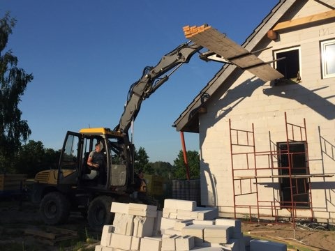 Żółto-szara koparka kołowa podnosi deski do okna w surowym stanie budynku z białego bloczka, obok rusztowanie i stos bloczków.