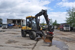 Żółto-czarna koparka kołowa Volvo na placu budowy, z łyżką skierowaną w dół, w tle budynek i inne pojazdy.