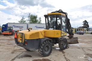 Żółta koparka kołowa z łyżką na tle błękitnego nieba z chmurami, zaparkowana na placu budowy. Widoczne opony z bieżnikiem i elementy hydrauliczne.