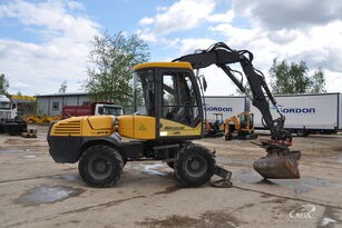 Żółta koparka kołowa Mecalac z łyżką, zaparkowana na placu budowy z widocznymi w tle drzewami i naczepą ciężarową.