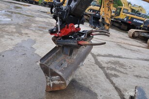 Szczegółowe ujęcie łyżki koparki z zamontowanym hydraulicznym chwytakiem, widoczne elementy mocujące w kolorze czerwonym, w tle zaparkowane maszyny budowlane.