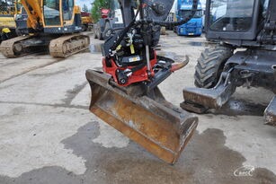 Łyżka koparki z hydraulicznym szybkozłączem, widoczne ślady użytkowania i błoto na łyżce, w tle inne maszyny budowlane i częściowo widoczna naczepa samochodu ciężarowego.