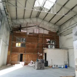 Wnętrze hali w trakcie prac izolacyjnych: ściana z ociepleniem z wełny mineralnej na rusztowaniu, widoczne okna, materiały budowlane i narzędzia na podłodze, pracownik w kasku na rusztowaniu.