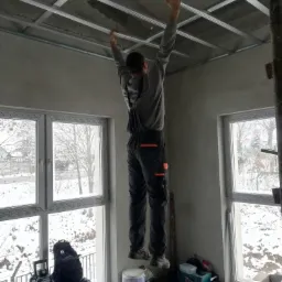 Monter zawiesza metalową konstrukcję sufitu podwieszanego w surowym wnętrzu, widok z dołu na tle okien z zimowym krajobrazem, narzędzia i sprzęt budowlany na podłodze.