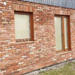 Murowana elewacja budynku z czerwonej cegły z widocznymi drewnianymi oknami i szarą drewnianą obudową, fragment dachu z blachy trapezowej, widoczne podpory na trawniku.