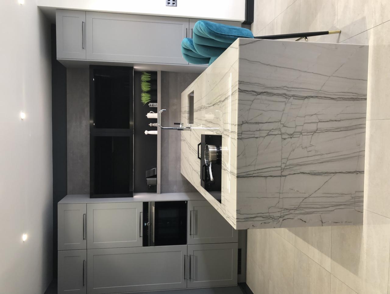 Szara kuchnia z wyspą z blatem z białego marmuru z szarymi żyłami, nowoczesny design, czarne AGD w zabudowie, turkusowe krzesło barowe.