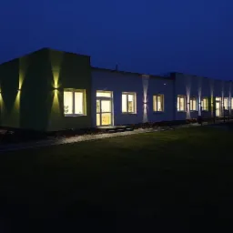 Oświetlony budynek z zewnątrz w nocy, z reflektorami punktowymi skierowanymi w dół, tworzącymi dekoracyjny efekt świetlny na elewacji.