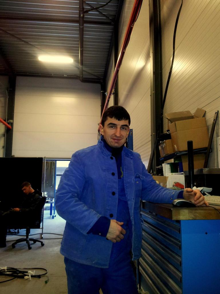 Mężczyzna w niebieskim kombinezonie roboczym, w warsztacie z blachy falistej, opierający się o niebieski stół warsztatowy. W tle drugi pracownik.