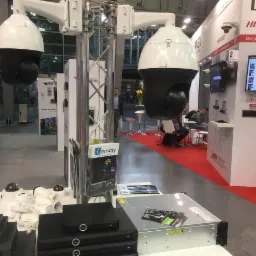 Ekspozycja systemów monitoringu na targach: dwie duże kamery PTZ na stalowych konstrukcjach, serwery i mniejsze kamery na białych stołach, czerwona wykładzina, logo Hikvision w tle.