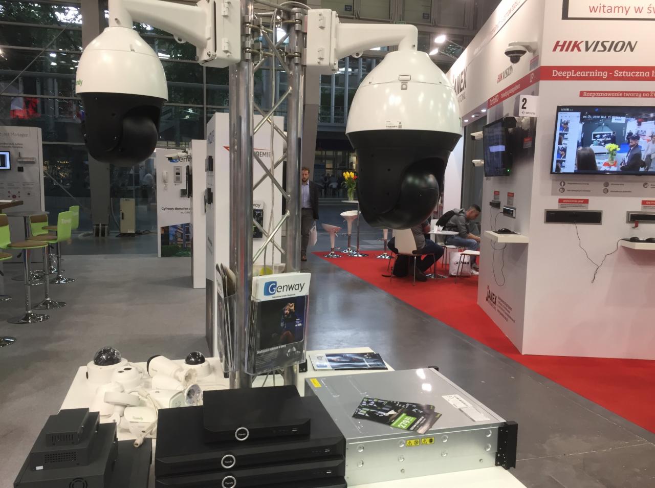 Ekspozycja systemów monitoringu na targach: dwie duże kamery PTZ na stalowych konstrukcjach, serwery i mniejsze kamery na białych stołach, czerwona wykładzina, logo Hikvision w tle.