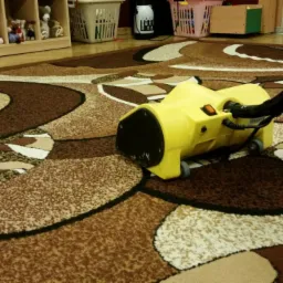 Żółty odkurzacz piorący Karcher na brązowym dywanie we wzory w pokoju dziecięcym z półkami pełnymi zabawek w tle.