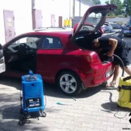 Mężczyzna w czarnej koszulce czyści wnętrze bagażnika czerwonego samochodu za pomocą żółtego odkurzacza piorącego. Obok stoi niebieski odkurzacz piorący, a drzwi samochodu są otwarte.