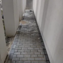 Ułożona ścieżka z szarej kostki betonowej, obrzeża z ciemniejszej kostki, pomiędzy budynkiem a kolumnami, w tle widoczny pył i narzędzia.