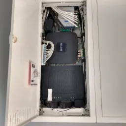 Szafka z centralą alarmową i routerem, widoczne kable, zasilacze i listwa zaciskowa, częściowo otwarta obudowa, logo BCS na urządzeniach i naklejce informacyjnej.