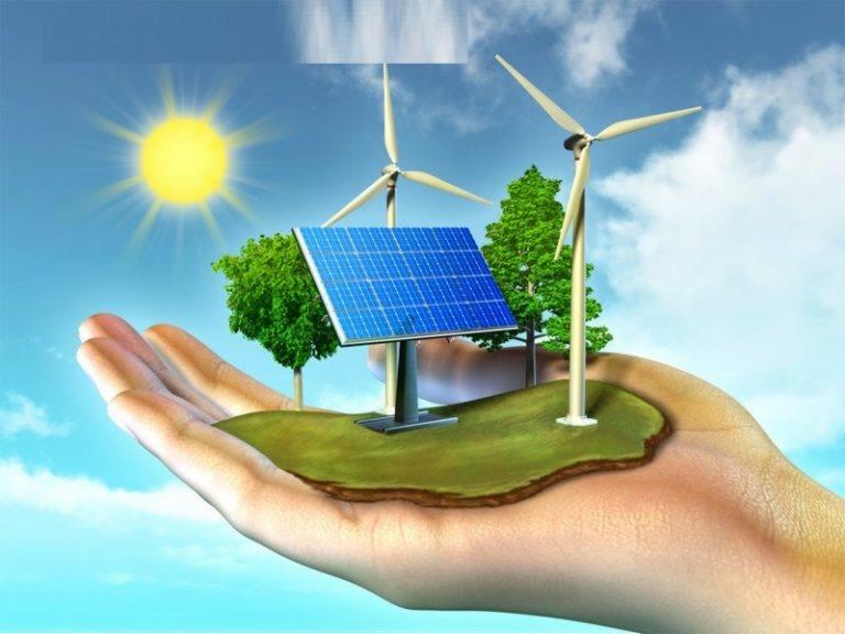 Dłoń trzymająca miniaturowy krajobraz z panelem fotowoltaicznym, turbinami wiatrowymi i drzewami na tle błękitnego nieba z słońcem.