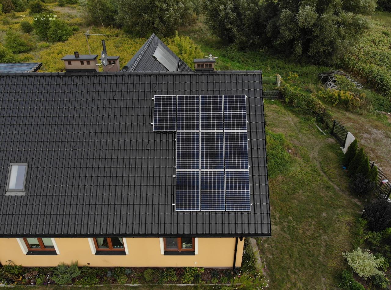 Widok z góry na dom z żółtą elewacją i ciemnym dachem, na którym zamontowano panele słoneczne w dwóch sekcjach; mniejsza sekcja z trzema panelami i większa z dziewięcioma, otoczone zielenią.