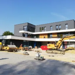 Żółty walec drogowy i koparka Liebherr na placu budowy przed nowoczesnym, szarym budynkiem z białymi elementami architektonicznymi i rusztowaniami w tle, w słoneczny dzień.