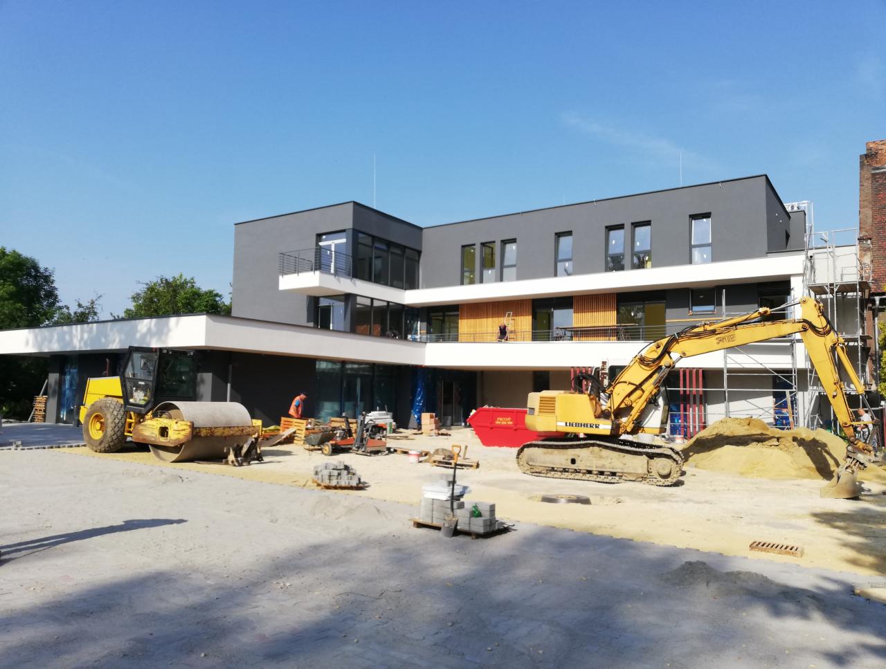 Żółty walec drogowy i koparka Liebherr na placu budowy przed nowoczesnym, szarym budynkiem z białymi elementami architektonicznymi i rusztowaniami w tle, w słoneczny dzień.