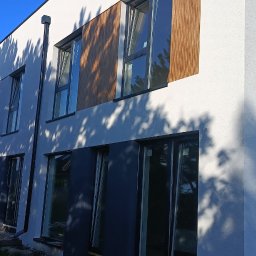 Usługi budowlane DAW-BUD - Nowoczesny budynek w trakcie wykończenia elewacji: biały tynk, drewniane panele, ciemne okna. Widoczne rusztowania i cień drzewa na fasadzie.