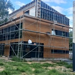 Usługi budowlane DAW-BUD - Dom w trakcie budowy z drewnianą elewacją i rusztowaniami. Widoczne okna z czarnymi ramami i fragment budynku z czerwonej cegły. Prace wykończeniowe w toku.