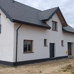 Usługi budowlane DAW-BUD - Nowoczesny dom jednorodzinny z ciemną dachówką i białymi ścianami. Widoczne okna z drewnianą ramą, metalowe balustrady, skrzynka gazowa. W tle inne domy.