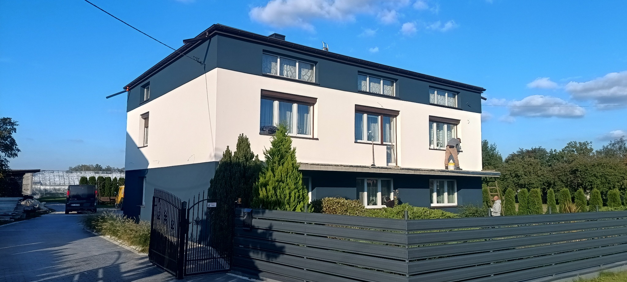 Dwukolorowy dom po renowacji elewacji. Pracownik maluje ścianę na biało, stojąc na dachu. Widoczny fragment ogrodzenia i zieleń. Słoneczny dzień.