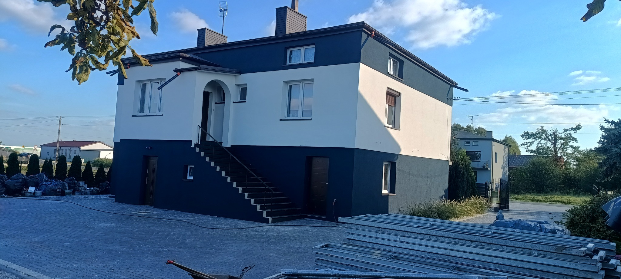 Dwukondygnacyjny dom po renowacji elewacji w kolorach bieli i granatu, z widocznymi schodami wejściowymi i stertą worków po farbie. Widok z dołu, słoneczny dzień.