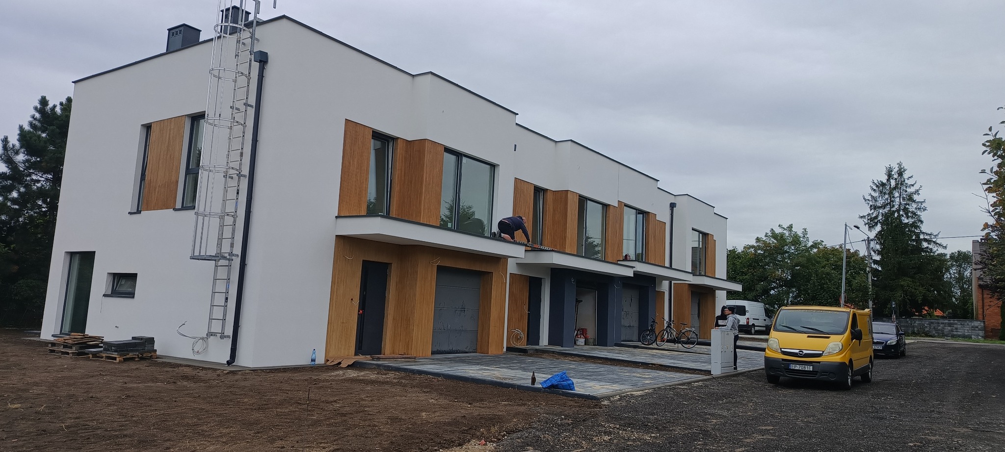 Nowoczesne domy w zabudowie szeregowej z drewnianą elewacją. Pracownik mierzy dach. Żółty van z logo firmy na podjeździe.