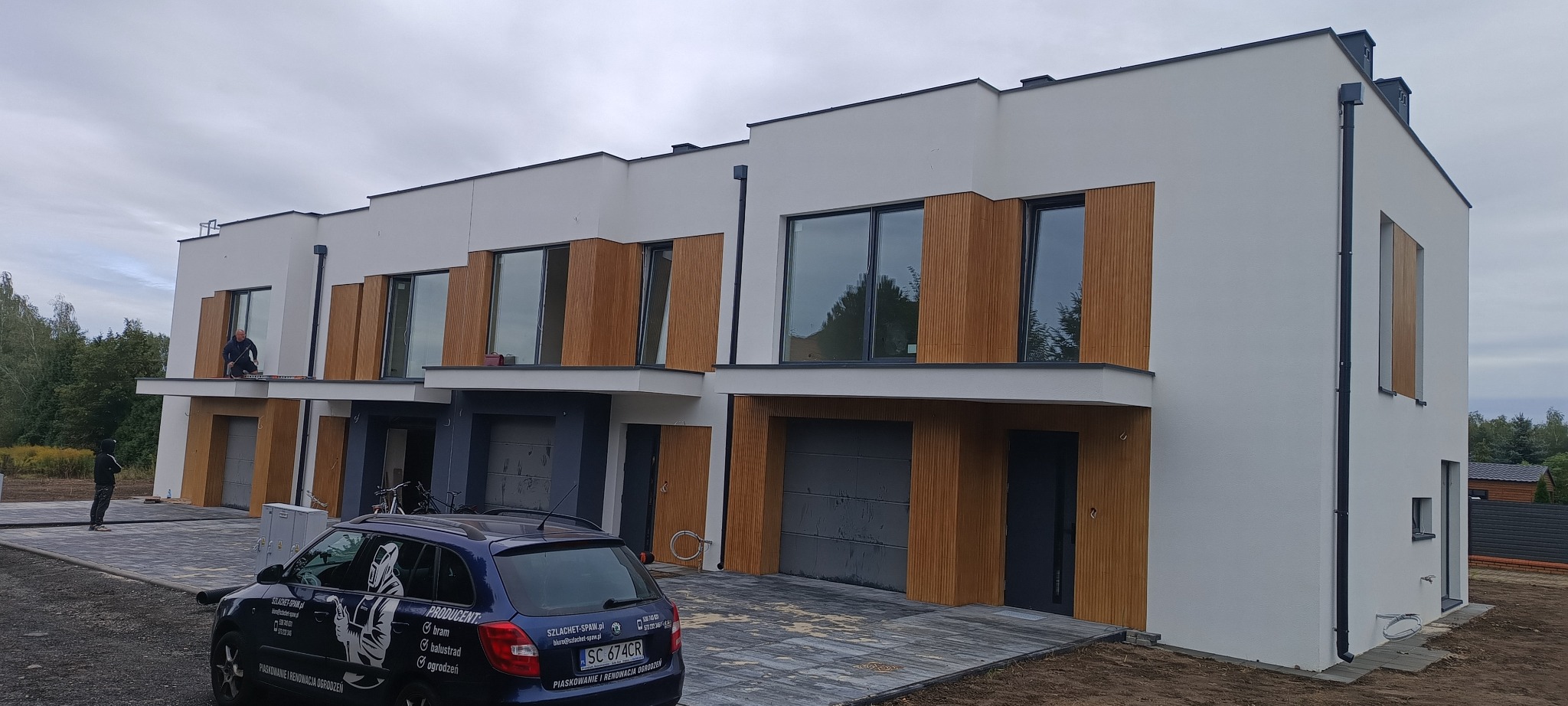 Nowoczesne domy szeregowe z drewnianą elewacją, pracownik na balkonie jednego z domów. Na pierwszym planie zaparkowany samochód z reklamą firmy.