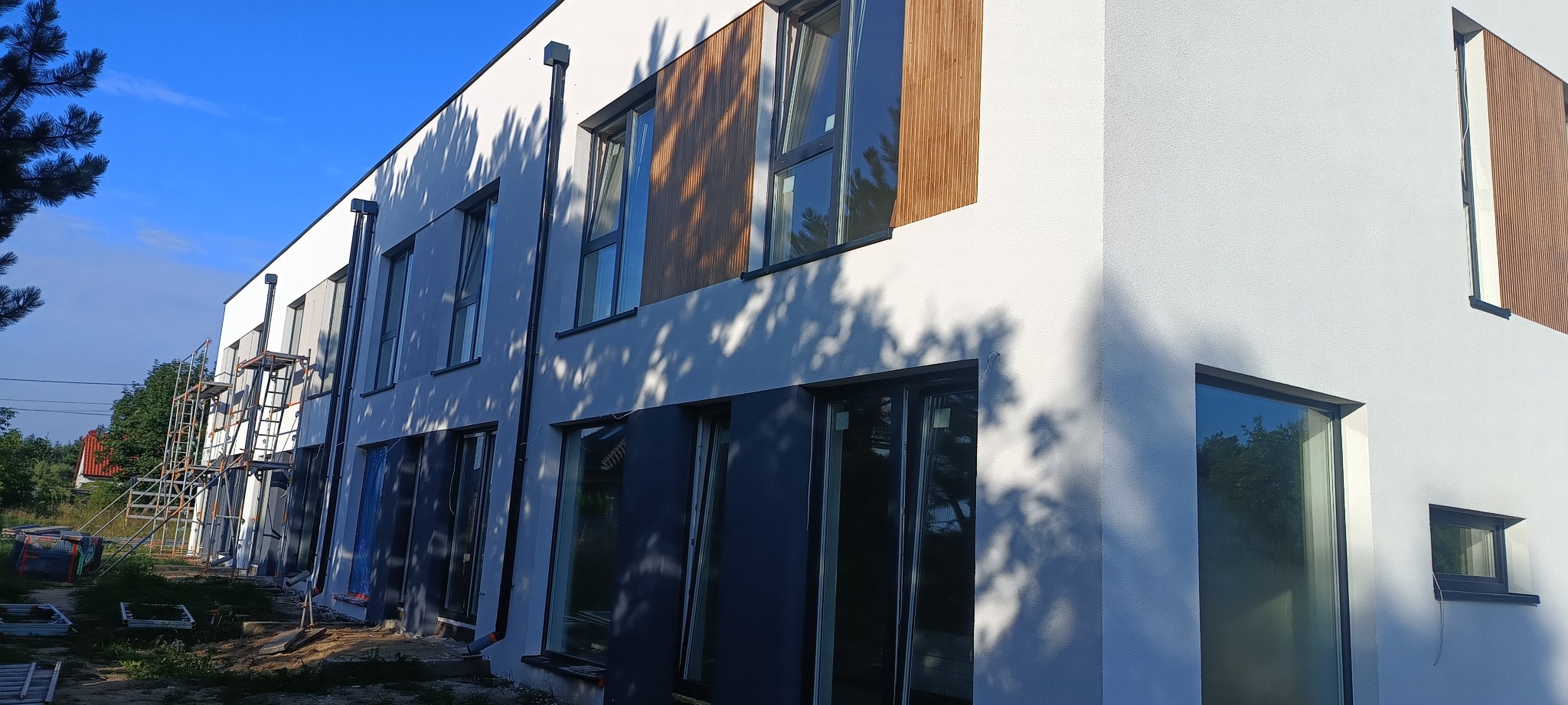 Nowoczesny budynek w trakcie wykończenia elewacji: biały tynk, drewniane panele, ciemne okna. Widoczne rusztowania i cień drzewa na fasadzie.
