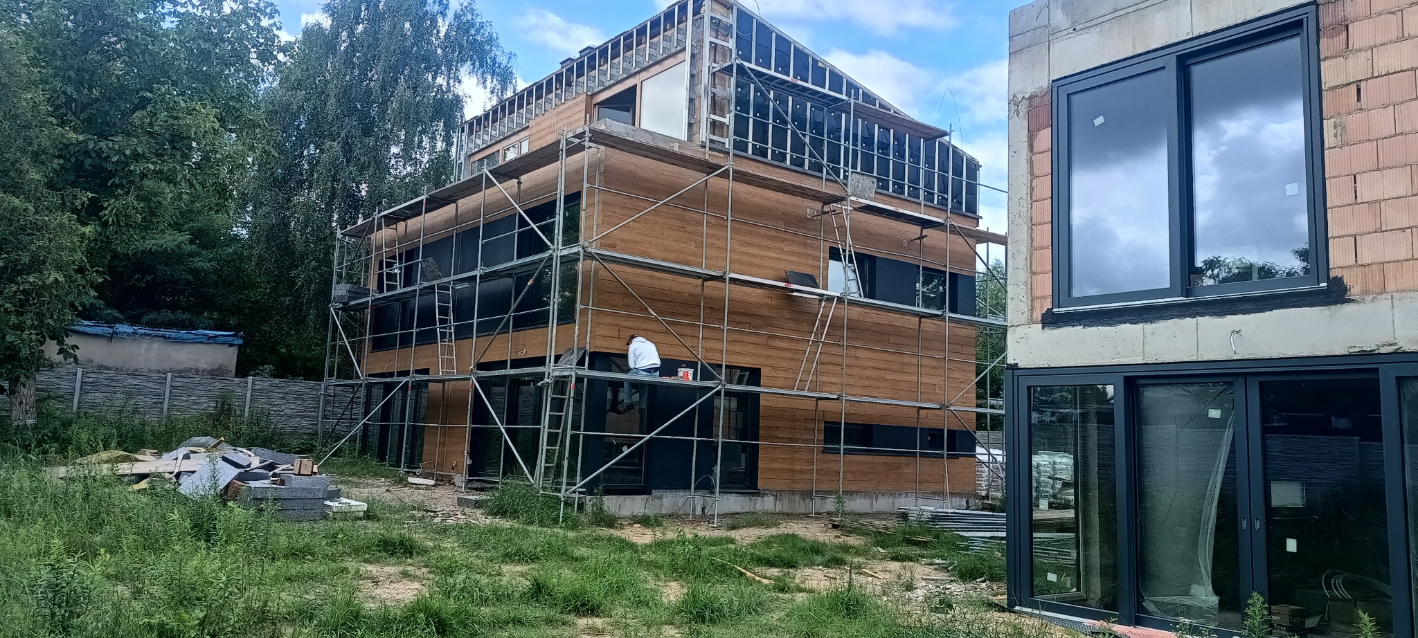 Dom w trakcie budowy z drewnianą elewacją i rusztowaniami. Widoczne okna z czarnymi ramami i fragment budynku z czerwonej cegły. Prace wykończeniowe w toku.