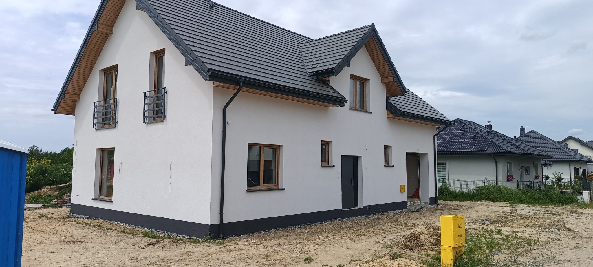 Nowoczesny dom jednorodzinny z ciemną dachówką i białymi ścianami. Widoczne okna z drewnianą ramą, metalowe balustrady, skrzynka gazowa. W tle inne domy.