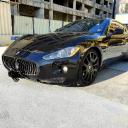 Błyszczący czarny Maserati GranTurismo z czarnymi felgami na tle miejskiego krajobrazu z budynkami w budowie i kontenerami.
