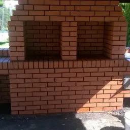 Nowo wymurowany, ceglany grill ogrodowy z wędzarnią, z widoczną metalową klapą i otworami wentylacyjnymi, usytuowany pod zadaszeniem z betonowym słupem, w tle zieleń ogrodu.