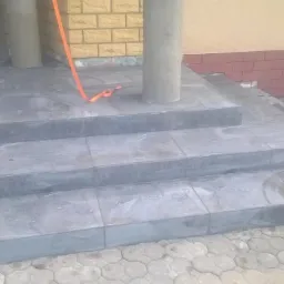 Wykończone, szare, ceramiczne schody wejściowe do budynku z kolumnami, widoczny fragment elewacji z cegły i wiadro po farbie.