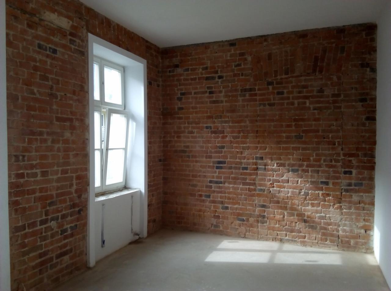 Surowe wnętrze z ceglanymi ścianami i nową wylewką podłogową, widoczne ślady po wcześniejszych instalacjach na ścianie, okno z białymi ramami.