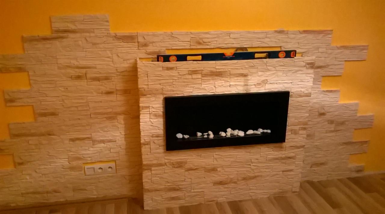 Ściana wykończona panelami imitującymi kamień, z wbudowanym czarnym kominkiem elektrycznym z białymi kamykami, poziomica widoczna w górnej części, żółta ściana w tle.