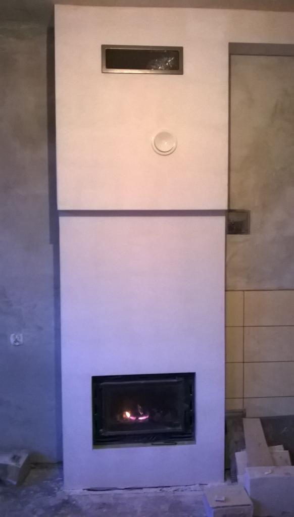 Wykończony kominek z wkładem, świeżo po nałożeniu gładzi, widoczne detale wykończeniowe i fragment ściany z płytkami.